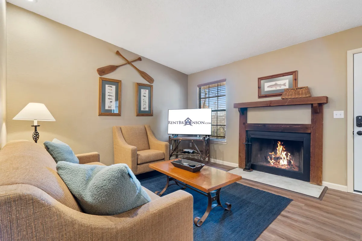 A cozy living room with a stone fireplace as its centerpiece, surrounded by a comfortable sofa and armchairs. Decorative oars hang on the wall, with a television nearby, and warm lighting enhances the inviting atmosphere. The space is designed for relaxation, whether socializing, watching TV, or enjoying a quiet evening by the fire.