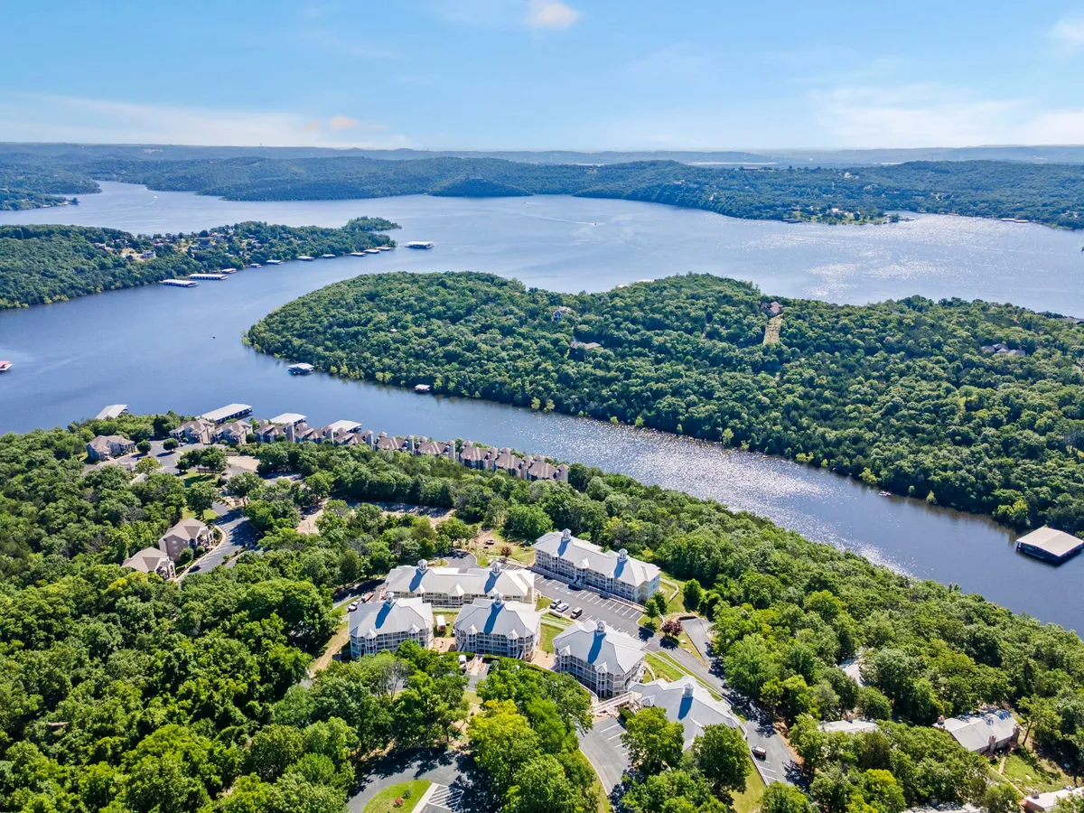 A wide, scenic view of lush greenery meeting sparkling water, with waterfront properties and forested hills in the background. The setting feels peaceful and expansive, inviting guests to enjoy outdoor activities by the lake.