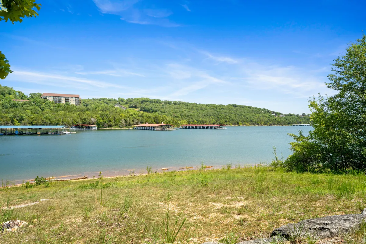 A sweeping view of a serene lake bordered by lush green hills, with distant buildings and docks dotting the landscape. The calm water and open sky evoke a sense of peace and natural beauty.