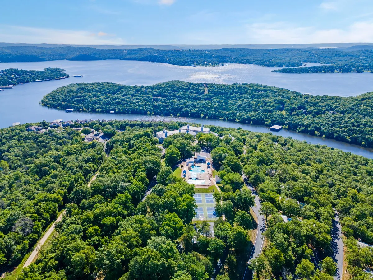 A vibrant exterior shot that captures a wide view of the resort community, featuring a sparkling swimming pool, well-maintained tennis courts, lush greenery, and a lake in the background. The scene evokes an active yet relaxed resort atmosphere, hinting at the variety of available activities.