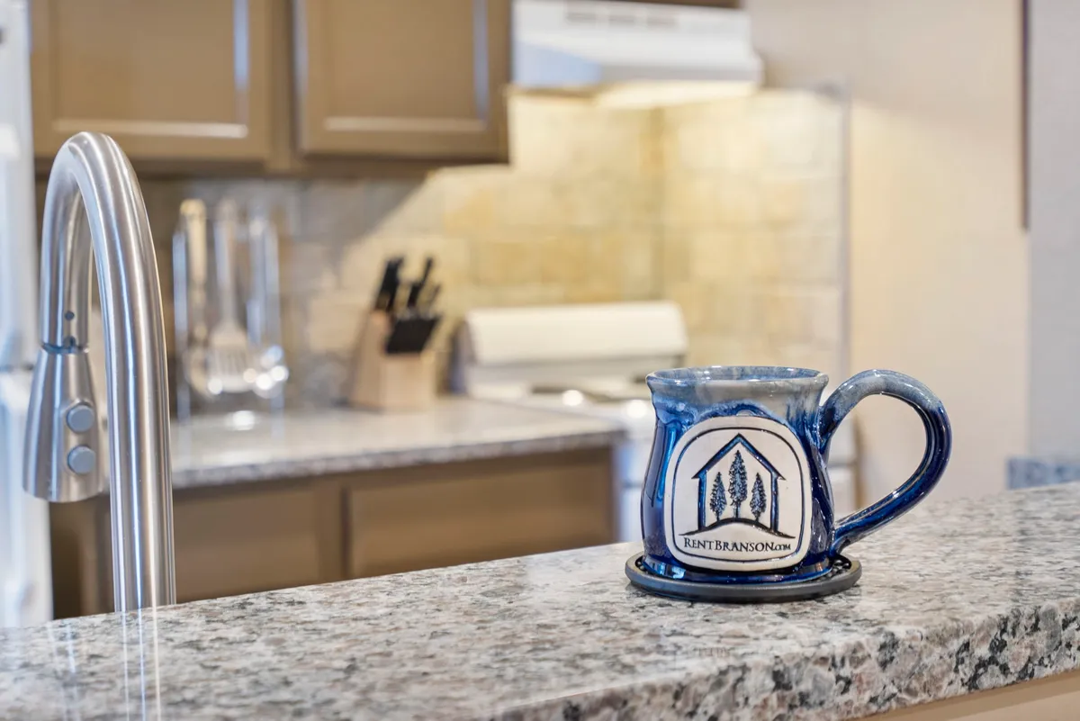 A cozy kitchen scene highlighting a ceramic mug resting on a granite countertop, with kitchen utensils and a stove in the background. The focus on a mug suggests morning rituals and comfort within the home setting.