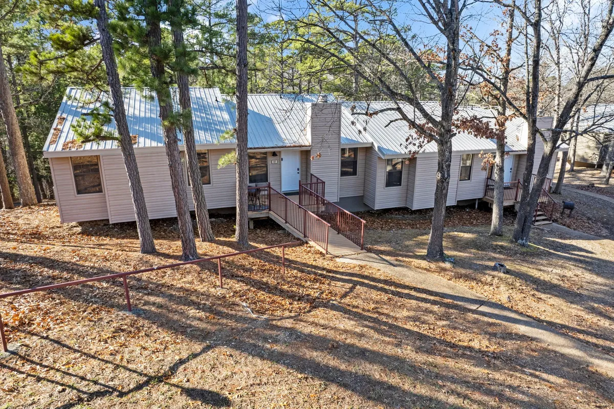 The image features the main entrance of a bungalow, with a ramp for easy access, surrounded by mature trees and a tranquil atmosphere. The metal roof and open porch invite guests to relax or prepare for a walk with their pet. The setting feels both welcoming and private, with natural shade and direct access to nature trails.