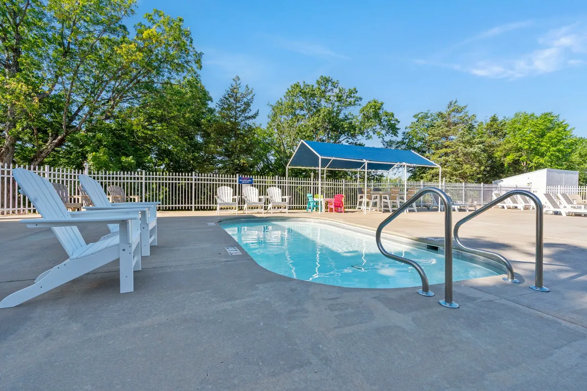 A sparkling seasonal outdoor pool bordered by lounge chairs and shaded by a canopy, set against a backdrop of mature green trees, offering space for swimming, relaxing, and socializing.